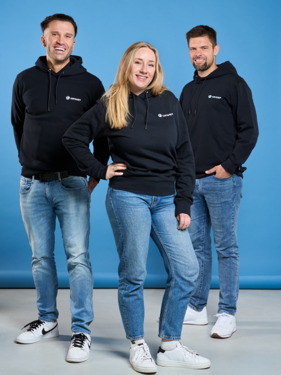Drei Models mit Jeanshosen und tragen Schwarzes Kapuzensweatshirt mit weißem TRILUX Logo auf der linken Brust